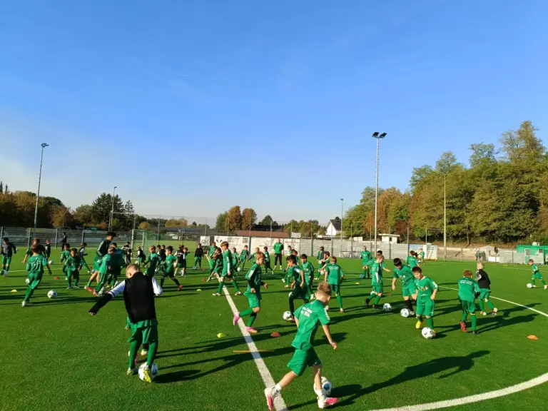 Viele Kinder in grünen Camp-Trikots dribbeln bei Sonnenschein mit Ball auf dem Kunstrasenplatz des FV09 Schwalbach