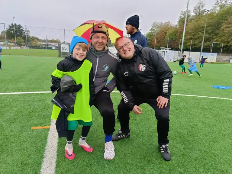 Zwei Trainer und ein lachender Teilnehmer posieren gemeinsam auf dem Platz, einer der Trainer trägt einen Regenschirmhut